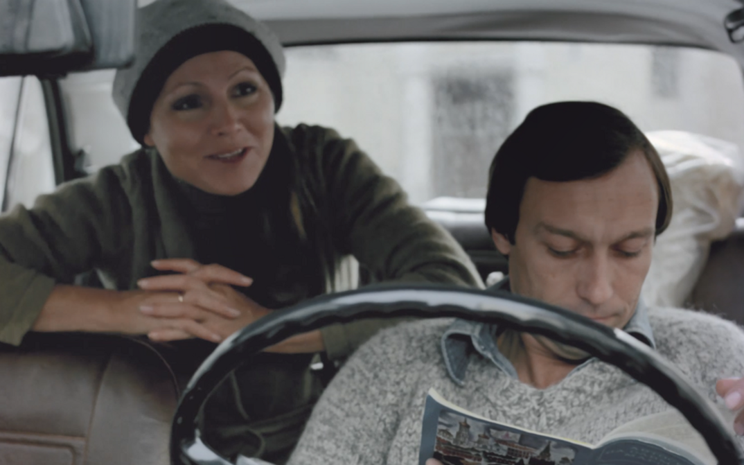 Film still from FLIGHTS IN DREAM AND REALITY: A man and a woman in a car, he sits in the front reading a magazine, she sits in the back leaning forward. 