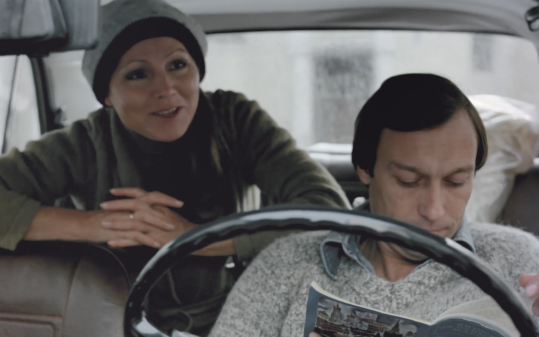 Filmstill aus TAGTRÄUMER: Ein Mann und eine Frau im Auto, er sitzt vorne und liest in einer Zeitschrift, sie sitzt hinten und beugt sich nach vorne.