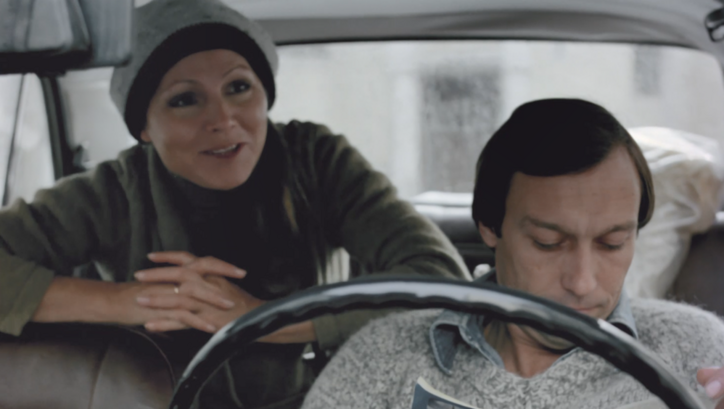 Filmstill aus TAGTRÄUMER: Ein Mann und eine Frau im Auto, er sitzt vorne und liest in einer Zeitschrift, sie sitzt hinten und beugt sich nach vorne. 