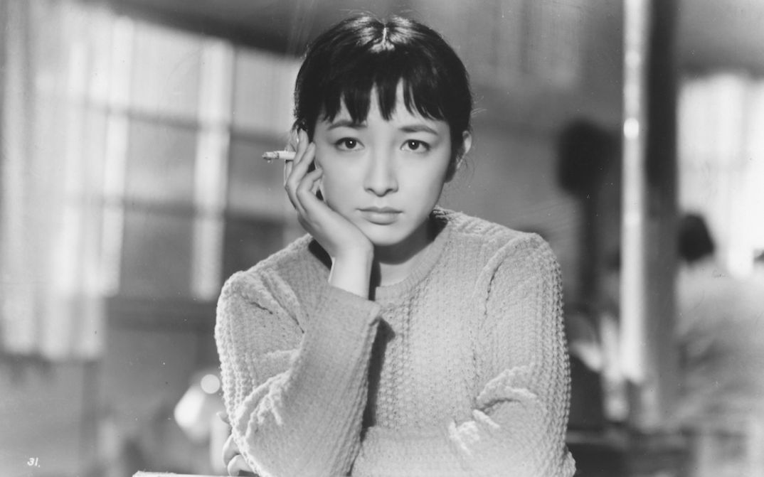 Filmstill aus TOKYO TWILIGHT: Eine junge Frau in einem Strickpulli sitzt an einem Tisch und blickt nachdenklich in die Kamera. Sie hat ihr Kinn auf die Hand gestützt und hält eine Zigarette.