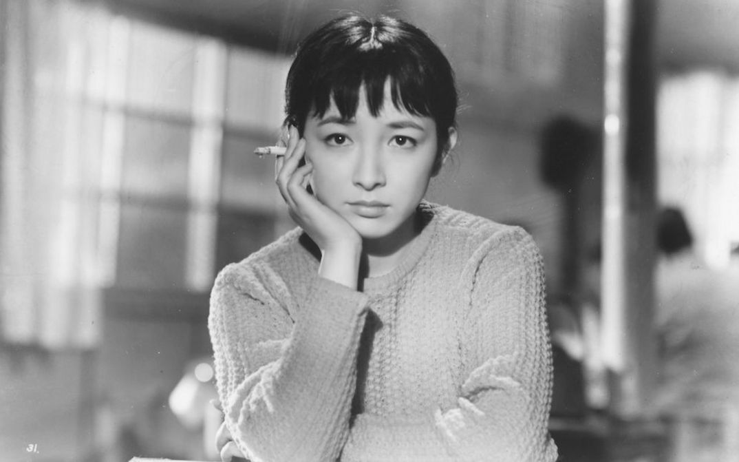 Filmstill aus TOKYO TWILIGHT: Eine junge Frau in einem Strickpulli sitzt an einem Tisch und blickt nachdenklich in die Kamera. Sie hat ihr Kinn auf die Hand gestützt und hält eine Zigarette.