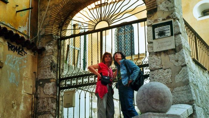 Film still from MIMI: Two women are standing on the street in front of an archway with a fence.