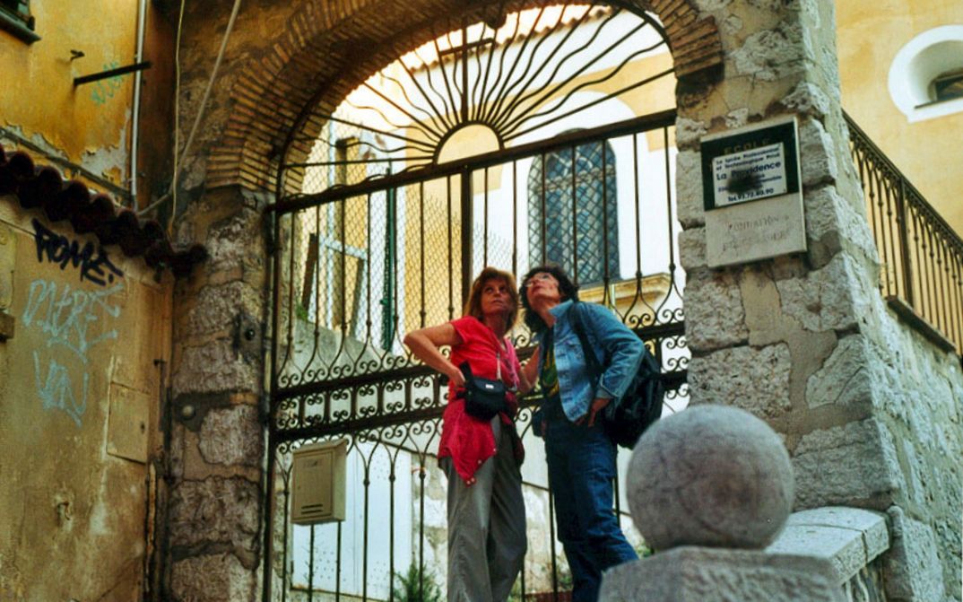 Film still from MIMI: Two women are standing on the street in front of an archway with a fence.