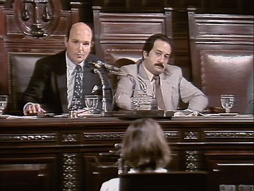 Filmstill aus dem Film "El juicio" von Ulises de la Orden. Zwei Männer in Anzügen sitzen auf einer Richterbank und blicken auf eine verschwommene Person im Vordergrund des Bildes.