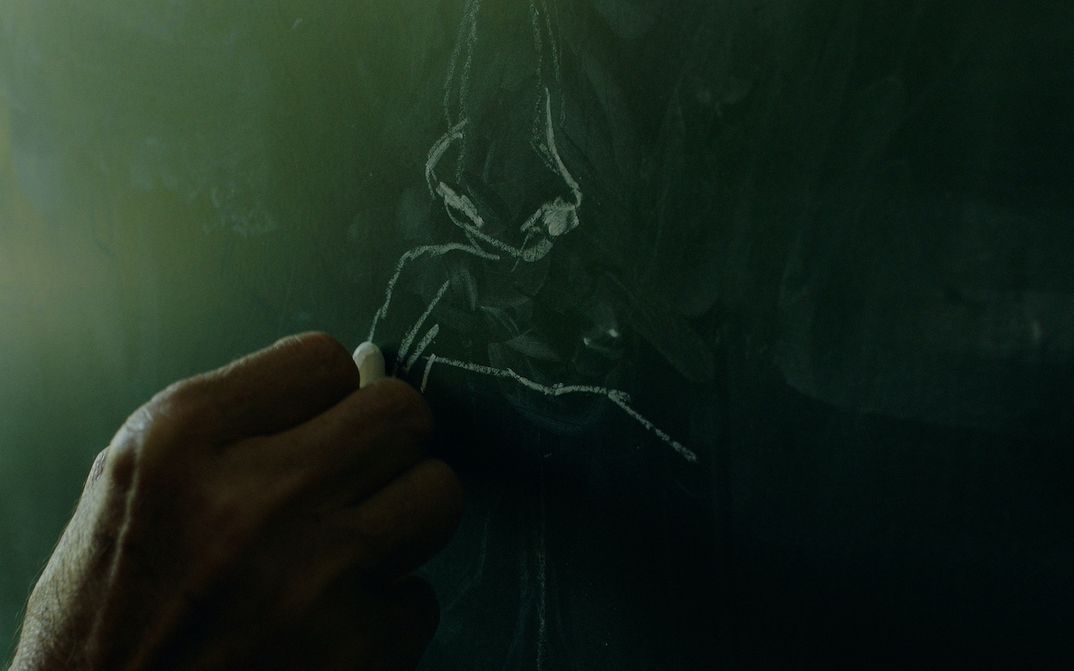 Filmstill aus I believe the portrait saved me: Eine Hand zeichnet mit einer Kreide auf eine Tafel.