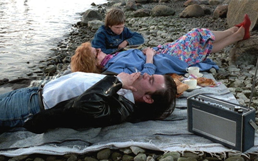 Filmstill aus ARIEL: Ein Paar und ein Kind liegen auf einer Picknickdecke an einem steinigen Strand.