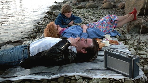 Film still from ARIEL: A couple and a child lie on a picnic blanket on a rocky beach.