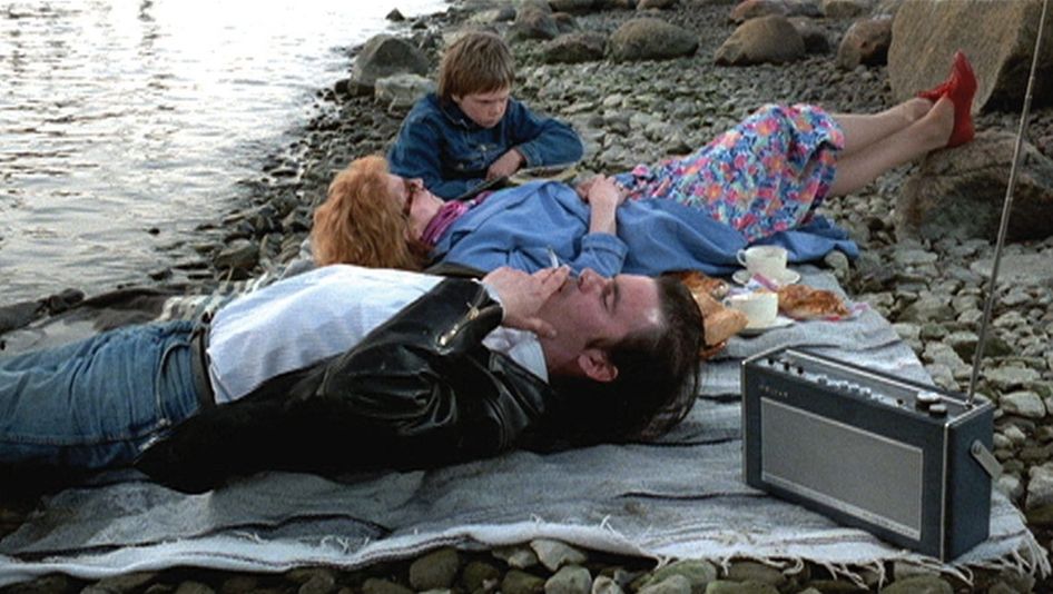 Film still from ARIEL: A couple and a child lie on a picnic blanket on a rocky beach.