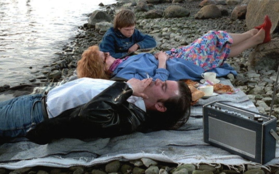 Filmstill aus ARIEL: Ein Paar und ein Kind liegen auf einer Picknickdecke an einem steinigen Strand.