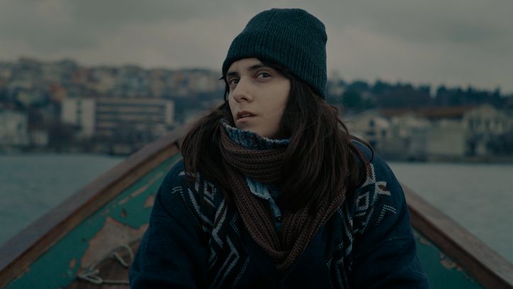 Filmstill aus TUZDAN KAIDE: Eine junge Frau in winterlicher Kleidung sitzt in einem kleinen Boot und blickt mit skeptischem Blick in die Kamera.