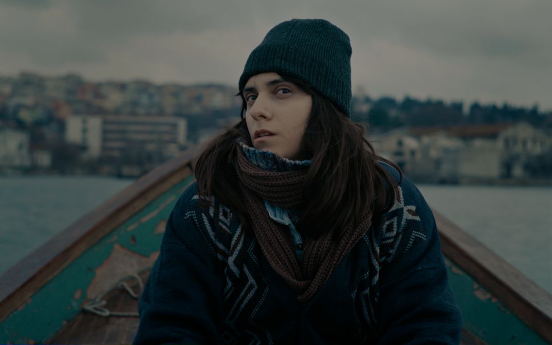 Filmstill aus TUZDAN KAIDE: Eine junge Frau in winterlicher Kleidung sitzt in einem kleinen Boot und blickt mit skeptischem Blick in die Kamera.