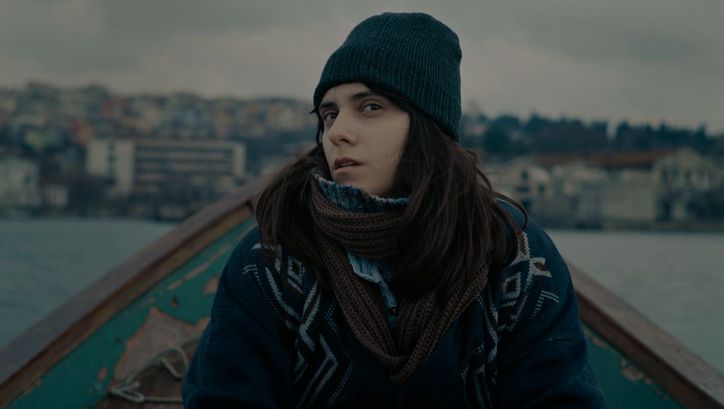 Filmstill aus TUZDAN KAIDE: Eine junge Frau in winterlicher Kleidung sitzt in einem kleinen Boot und blickt mit skeptischem Blick in die Kamera.