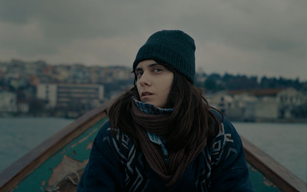 Filmstill aus TUZDAN KAIDE: Eine junge Frau in winterlicher Kleidung sitzt in einem kleinen Boot und blickt mit skeptischem Blick in die Kamera.