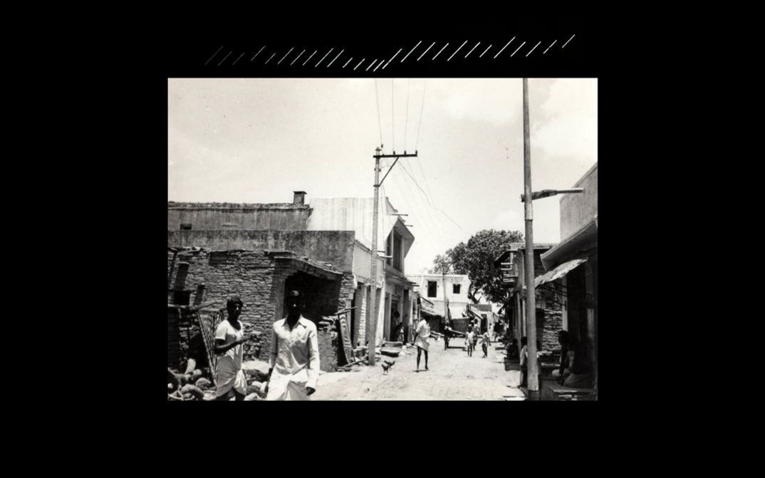 Filmstill aus dem Film „beneath the placid lake“ von Kush Badhwar und Vyjayanthi Rao. Auf schwarzem Hintergrundwird ein Schwarz-Weiß-Film auf eine Leinwand projiziert, der eine belebte Straße in einem Arbeiterviertel zeigt, gesäumt von Häusern.