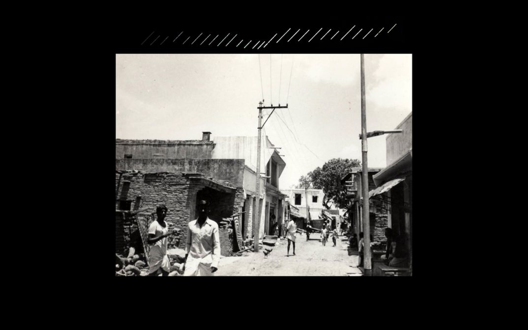 Filmstill aus dem Film „beneath the placid lake“ von Kush Badhwar und Vyjayanthi Rao. Auf schwarzem Hintergrundwird ein Schwarz-Weiß-Film auf eine Leinwand projiziert, der eine belebte Straße in einem Arbeiterviertel zeigt, gesäumt von Häusern.