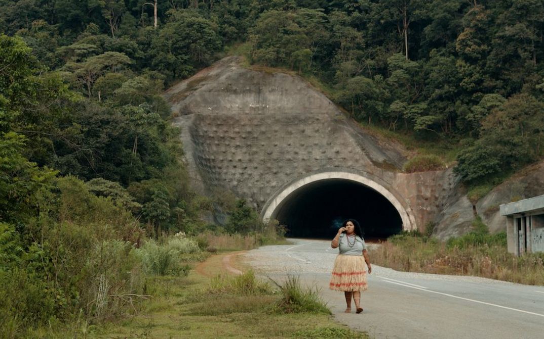 Film still from O ESTRANHO: A woman walks along a street. Behind her you can see the entrance to a tunnel, the road is lined with bushes.