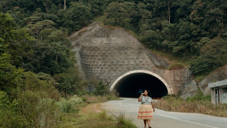 Film still from O ESTRANHO: A woman walks along a street. Behind her you can see the entrance to a tunnel, the road is lined with bushes.