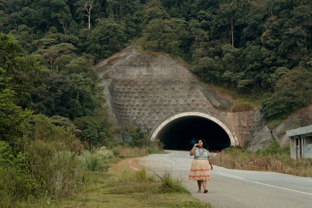 Filmstill aus O ESTRANHO: Eine Frau geht eine Straße entlang. Hinter ihr sieht man den Eingang zu einem Tunnel, die Straße ist gesäumt von Gebüsch.