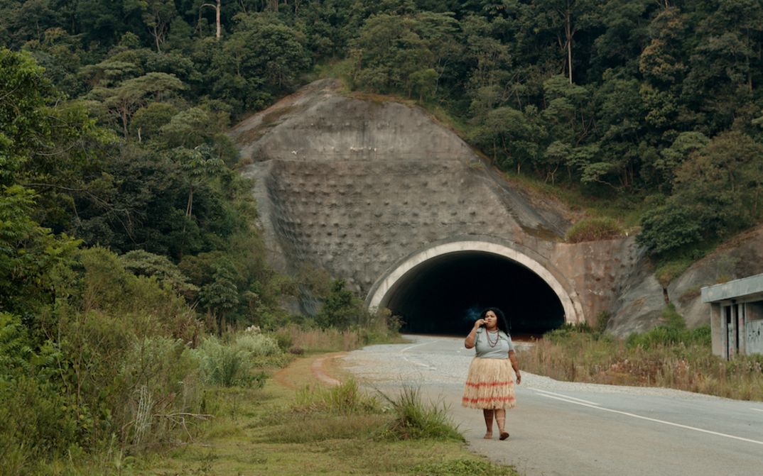 Filmstill aus O ESTRANHO: Eine Frau geht eine Straße entlang. Hinter ihr sieht man den Eingang zu einem Tunnel, die Straße ist gesäumt von Gebüsch.