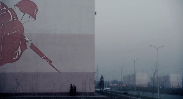 Filmstill aus CADET von Adilkhan Yerzhanov. Das Bild zeigt eine Hauswand, Straße und Gebäude im Hintergund. Auf der Hauswand ist ein Soldat mit Waffe gemalt. 