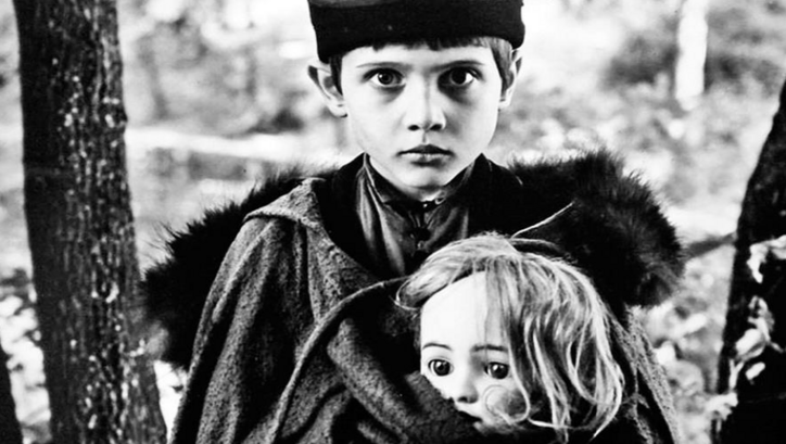Filmstill aus AMONG GREY STONES: Ein Junge hält eine Puppe im Arm und blickt in die Kamera.
