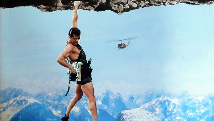 Film still from CLIFFHANGER: A climber is hanging from a rock by one hand and looking down into the abyss. A helicopter can be seen in the background.