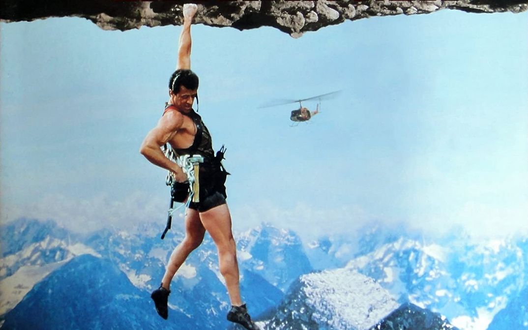 Filmstill aus CLIFFHANGER: Ein Kletterer hängt an einer Hand an einem Felsen und blickt in den Abgrund. Im Hintergrund ist ein Hubschrauber zu sehen.