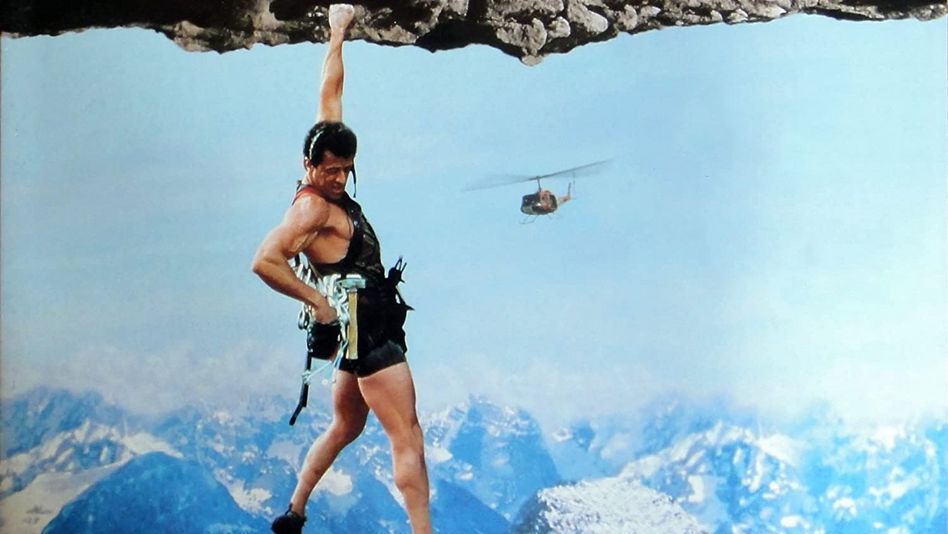Filmstill aus CLIFFHANGER: Ein Kletterer hängt an einer Hand an einem Felsen und blickt in den Abgrund. Im Hintergrund ist ein Hubschrauber zu sehen.