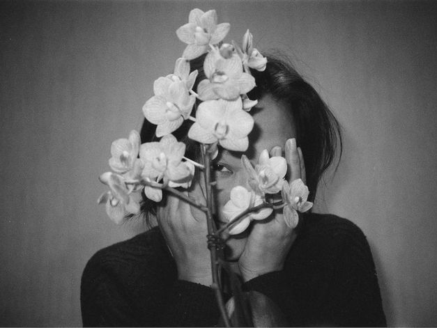Filmstill aus JET LAG: Eine junge Frau blickt hinter einer Blume hervor, von der sie halb verdeckt wird.