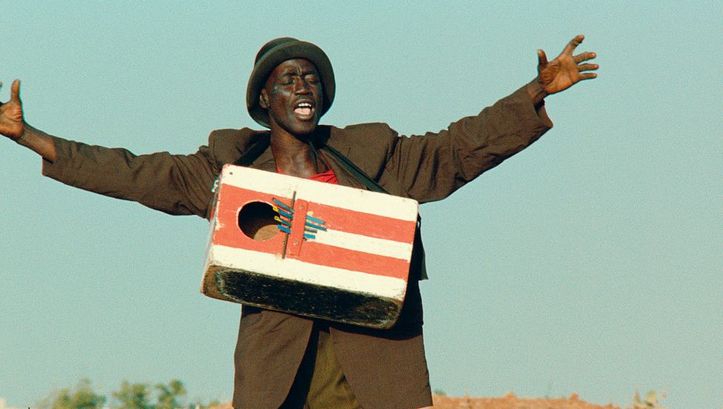 Film still from LE FRANC: A man stands in the landscape with his arms outstretched and a painted wooden box hanging around his neck.