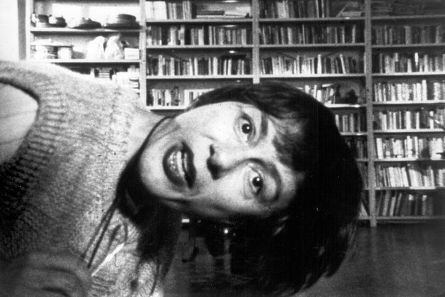 Film still from Yvonne Rainer’s „The Man Who Envied Women“. A woman leans into the picture from the left side and looks frontally at the camera. In the background a bookshelf.