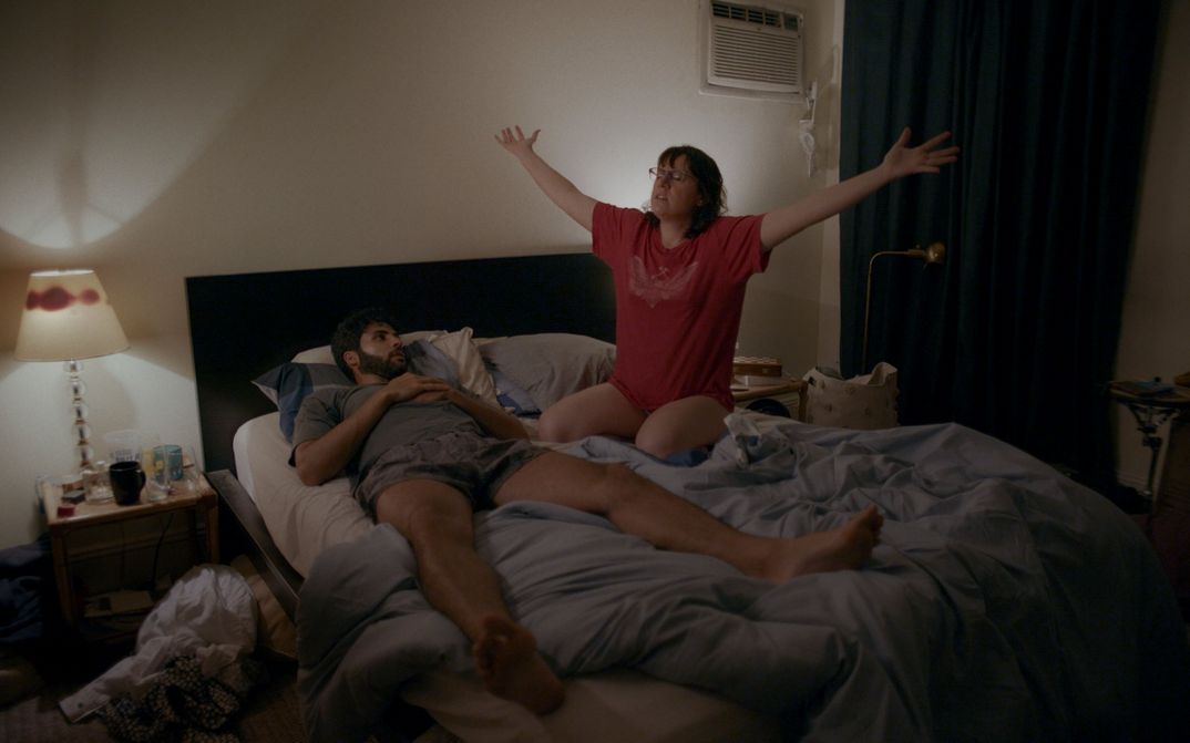 Film still from THE FEELING THAT THE TIME FOR DOING SOMETHING HAS PASSED: A man and a woman in an unmade bed. She is sitting and holding her arms outstretched.