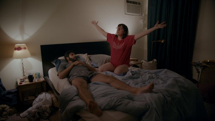 Filmstill aus THE FEELING THAT THE TIME FOR DOING SOMETHING HAS PASSED: Ein Mann und eine Frau in einem ungemachen Bett. Sie sitzt und hält die Arme ausgestreckt.