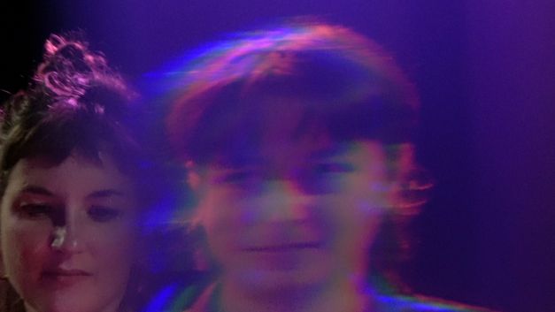 Film still from Stéphanie Lagarde’s film “Extra Life (And Decay)”. A blurred face occupies the foreground, while a second one appears clearly in the background, with colored reflections dancing on the features of the first.