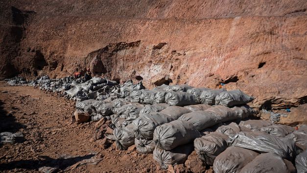 Film still from Petna Ndaliko Katondolo’s film “Mikuba”. At the foot of an arid mountain, rows of filled plastic bags are piled up, with men bustling between them in the distance.