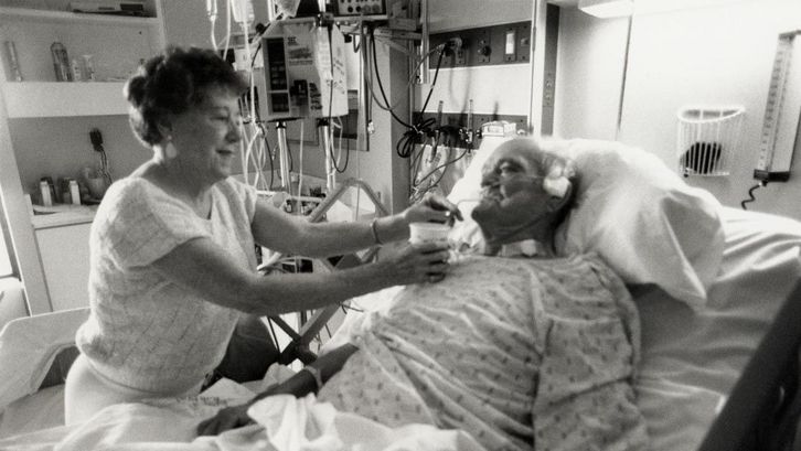 Film still from NEAR DEATH. A man lies in a hospital bed, a woman gives him something to drink.