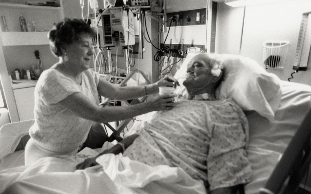 Film still from NEAR DEATH. A man lies in a hospital bed, a woman gives him something to drink.