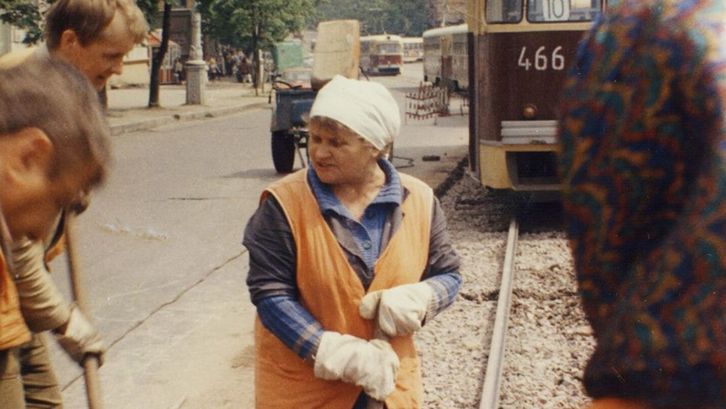 Filmstill aus ORANGENE WESTEN: Einige Straßenarbeiterinnen und -arbeiter vor einer Straßenbahn. In der Mitte eine Frau in oranger Weste.