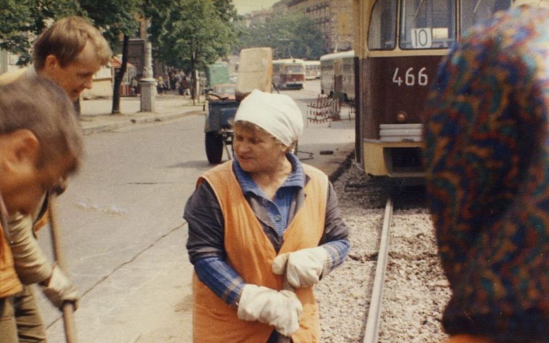 Filmstill aus ORANGENE WESTEN: Einige Straßenarbeiterinnen und -arbeiter vor einer Straßenbahn. In der Mitte eine Frau in oranger Weste.