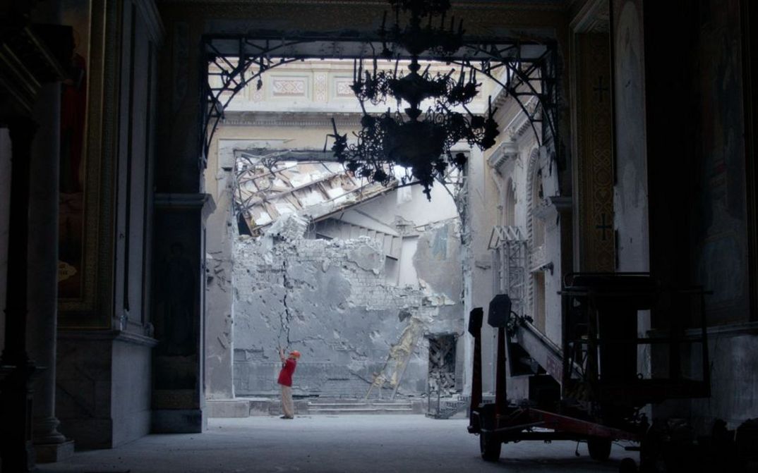 We are standing in a passageway and can see a damaged wall in the background. A man is standing at the back taking photographs.
