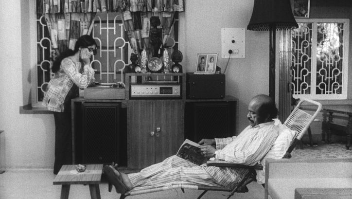 Film still from THAMP. A man and a woman in a living room, he is sitting in a deck chair reading, she is standing at the record player listening to the music.