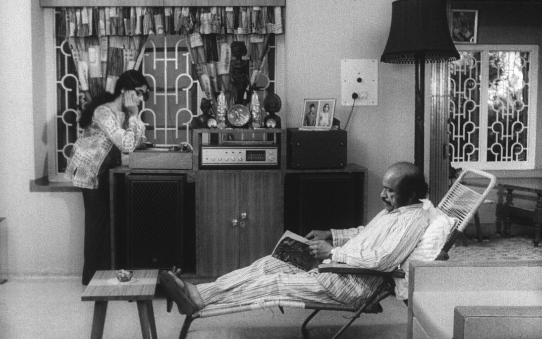 Filmstill aus THAMP. Ein Mann und eine Frau in einem Wohnzimmer, er sitzt in einem Liegestuhl und liest, sie steht am Plattenspieler und hört der Musik zu.