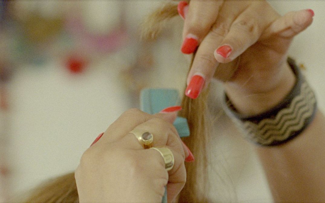 Still from the film "Mis Dos Voces" by Lina Rodriguez. We see a close-up of two hands with red painted fingernails running a blue comb through blonde hair.