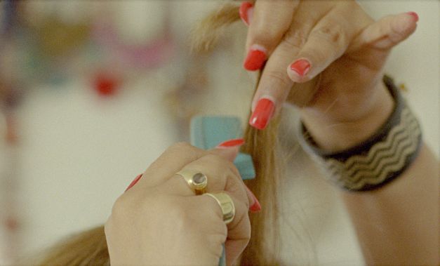 Filmstill aus „My Two Voices“ von Lina Rodriguez. Nahaufnahme von zwei Händen mit rot lackierten Fingernägeln. Die eine Hand hält blondes Haar in die Höhe, die andere Hand fährt mit einem blauen Kamm durch das Haar.