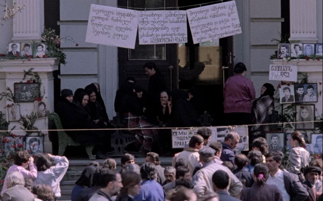 A protest scene: a crowd in front of a building, with placards and photos of faces.