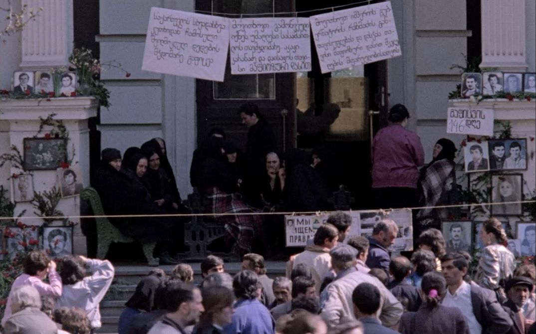 Eine Protestszene: Eine Menschenmenge vor einem Gebäude, mit Plakaten und Fotos von Gesichtern. 