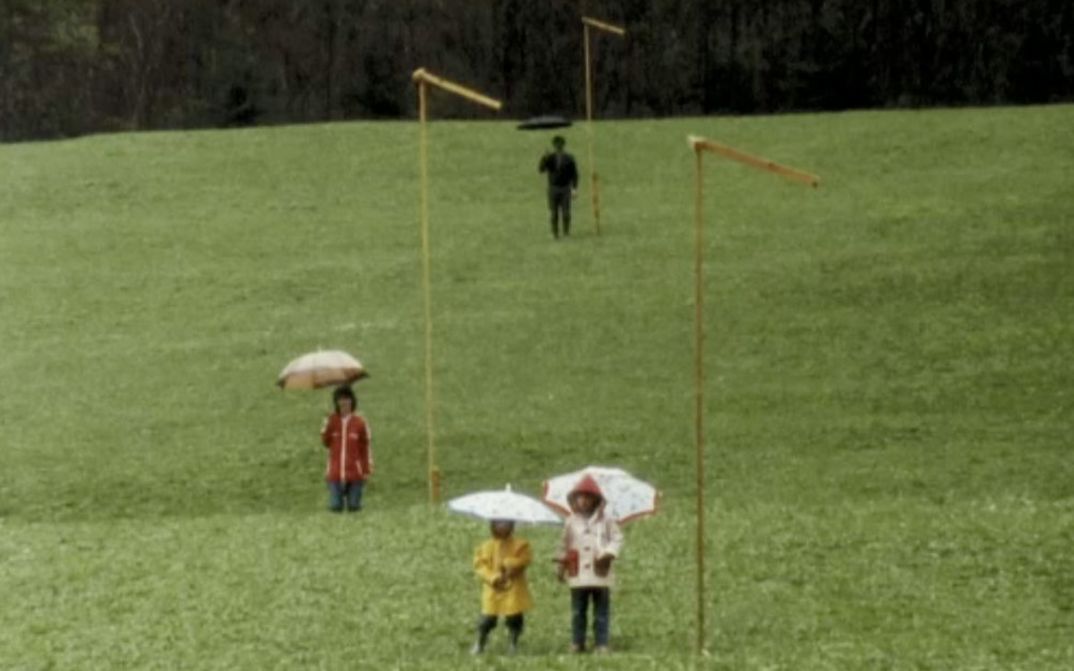 Filmstill aus DER GRÜNE BERG: Vier Menschen stehen verteilt und mit Regenschirmen auf einer Wiese.