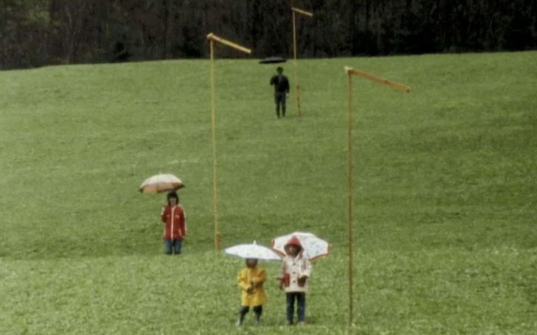 Film still from DER GRÜNE BERG: Four people are standing spread out and with umbrellas on a meadow.