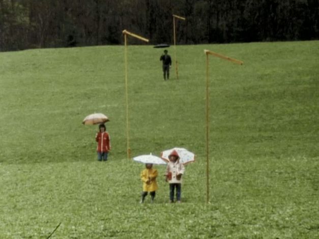 Filmstill aus DER GRÜNE BERG: Vier Menschen stehen verteilt und mit Regenschirmen auf einer Wiese.