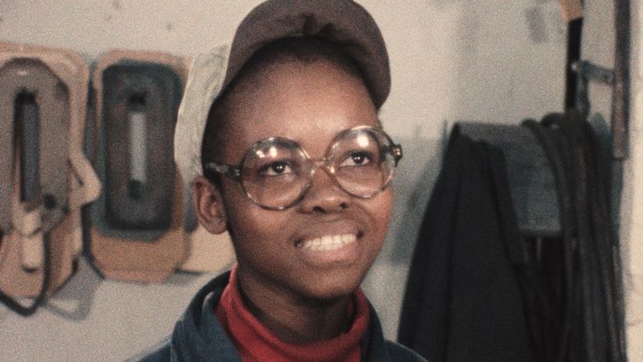 Film still from GLÜCK AUF – BILDER EINER BEGEGNUNG: A young person with a white cap and round glasses is grinning at the camera.